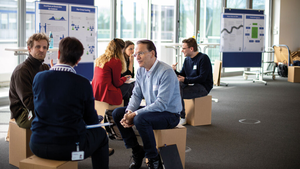 Workshop-Situation mit mehreren Kleingruppen, die auf Hockern im Stuhlkreis sitzen und intensiv diskutieren, im Hintergrund sind Moderationsposter sichtbar.