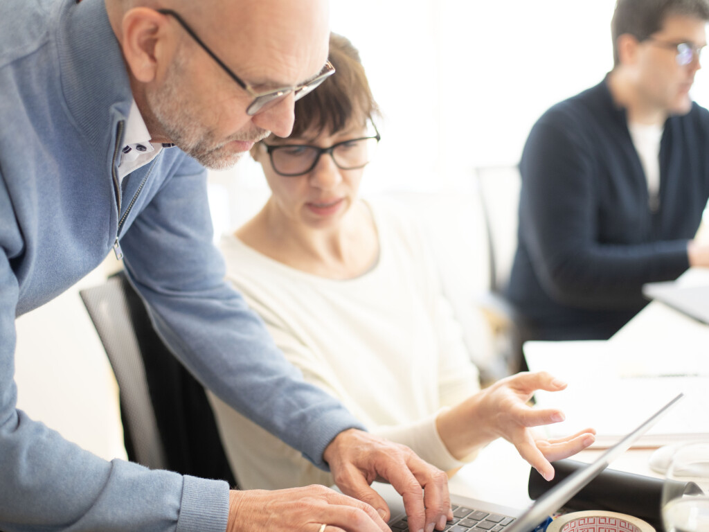 Trainer unterstützt Teilnehmerin am Laptop während einer Schulung, weitere Person arbeitet im Hintergrund.
