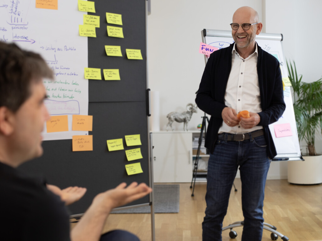 Trainer leitet Workshop vor Pinnwand mit bunten Moderationskarten, Teilnehmer im Gespräch sichtbar.