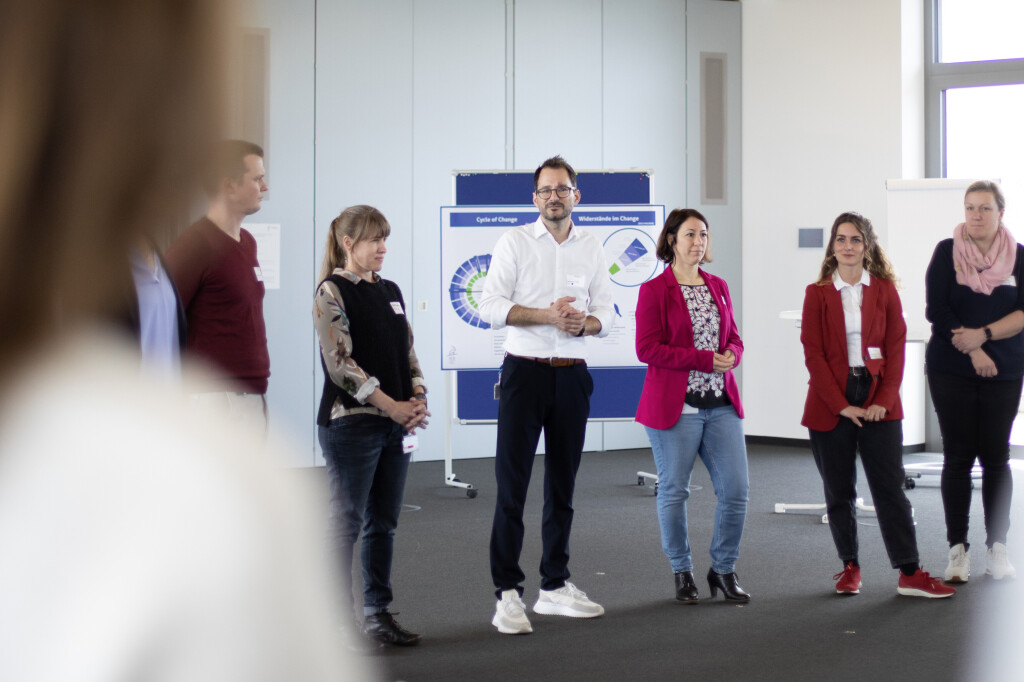 Teilnehmende stehen im Kreis während eines Change-Workshops, im Hintergrund Visualisierungen zu Veränderungsprozessen.