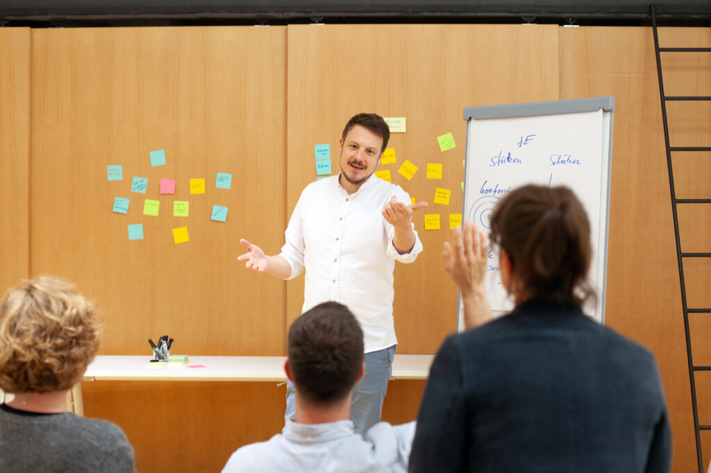 Trainer erklärt Moderationstechniken im Workshop, bunte Haftnotizen an der Wand, Teilnehmende stellen Fragen.