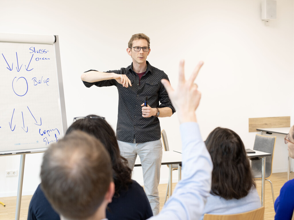 Trainer erklärt Inhalte an einem Flipchart während eines Workshops; Teilnehmende hören zu, eine Person meldet sich – interaktives Coaching und Training.