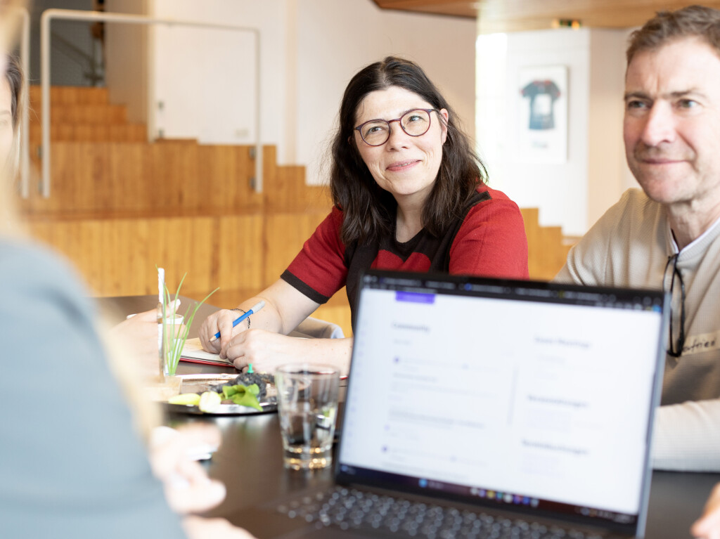 Teilnehmerinnen und Teilnehmer sitzen am Tisch in einem Workshop, lächeln und machen sich Notizen; im Vordergrund ein Laptop – Teamarbeit, Coaching und Zusammenarbeit.