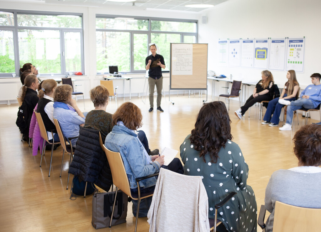 Fortgeschrittenes Präsentationstraining im Seminarraum: Trainer spricht zu Gruppe im Stuhlkreis, Fokus auf Wirkung & professionellem Auftritt.“.