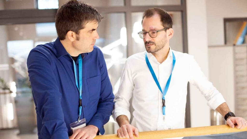 Zwei Männer mit Namensschildern im Gespräch in einem modernen Büro, beide tragen blaue Schlüsselbänder und wirken konzentriert und engagiert.