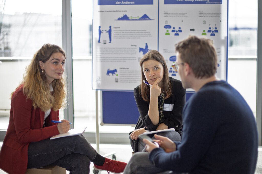 Drei junge Erwachsene sitzen in einer Diskussionsrunde, halten Stifte und Notizblöcke, und sprechen konzentriert miteinander; im Hintergrund Poster mit Coaching-Inhalten.