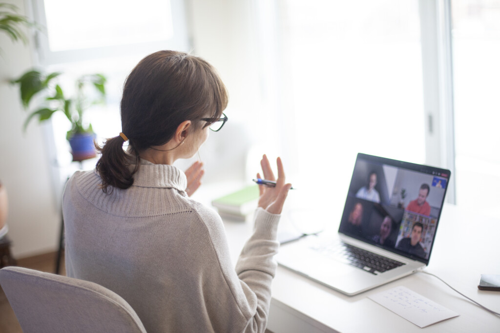 Frau mit Brille sitzt am Schreibtisch und nimmt an einem virtuellen Meeting auf einem Laptop mit sechs weiteren Personen teil.