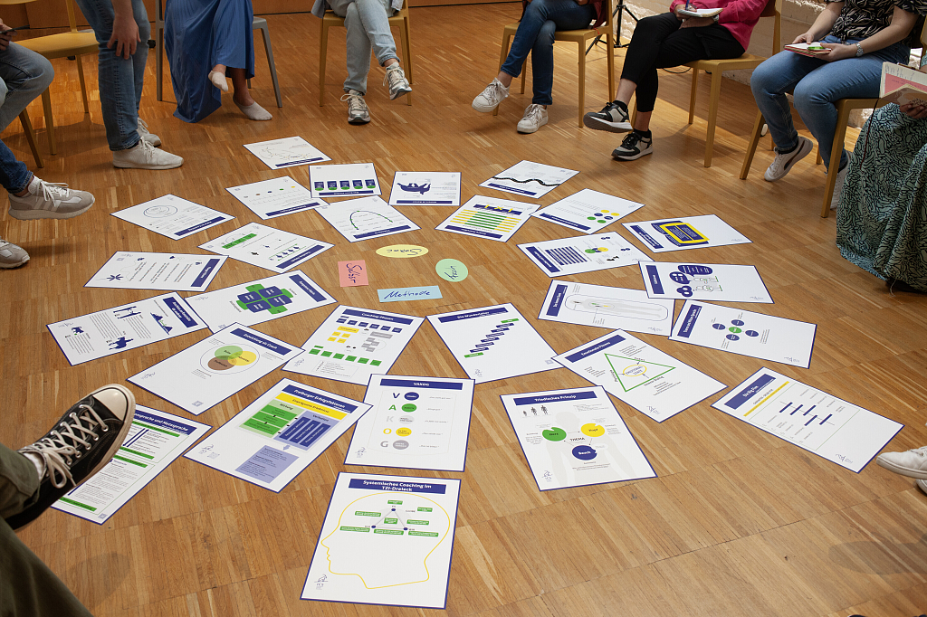 Teilnehmende sitzen im Kreis, Coaching-Methodenkarten und Modelle liegen auf dem Boden – gemeinsames Arbeiten an Konzepten und Prozessen.