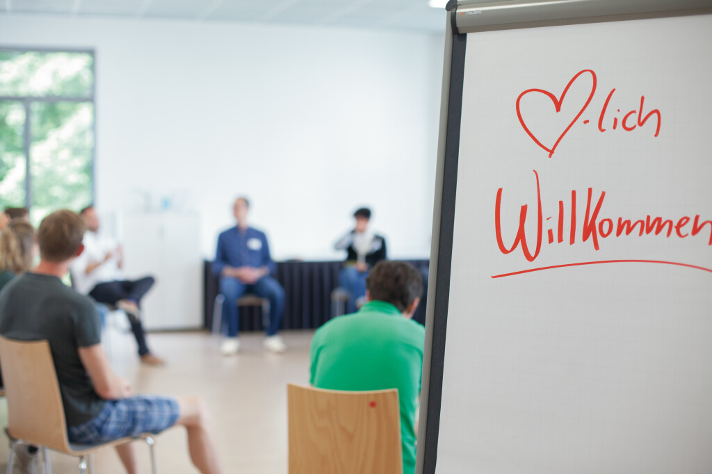 Flipchart mit der handschriftlichen Begrüßung „Herzlich Willkommen“ im Vordergrund, im Hintergrund eine sitzende Gruppe in einem Seminarsetting.
