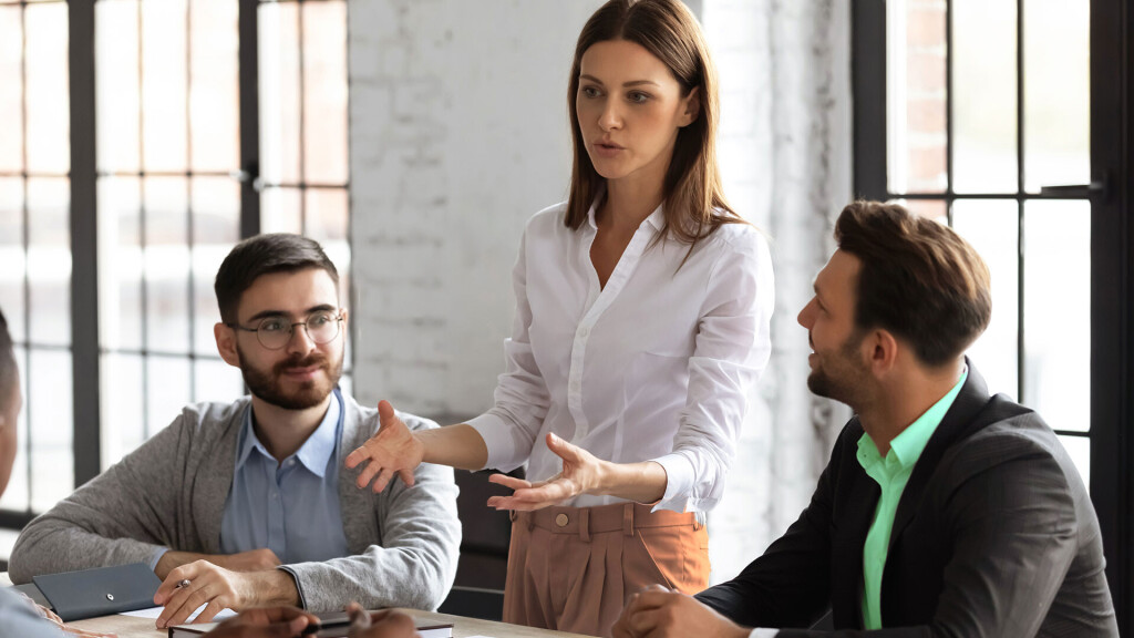 Frau in weißer Bluse spricht gestikulierend in einem Meeting, während zwei Männer aufmerksam zuhören – moderne Büroumgebung mit großen Fenstern.
