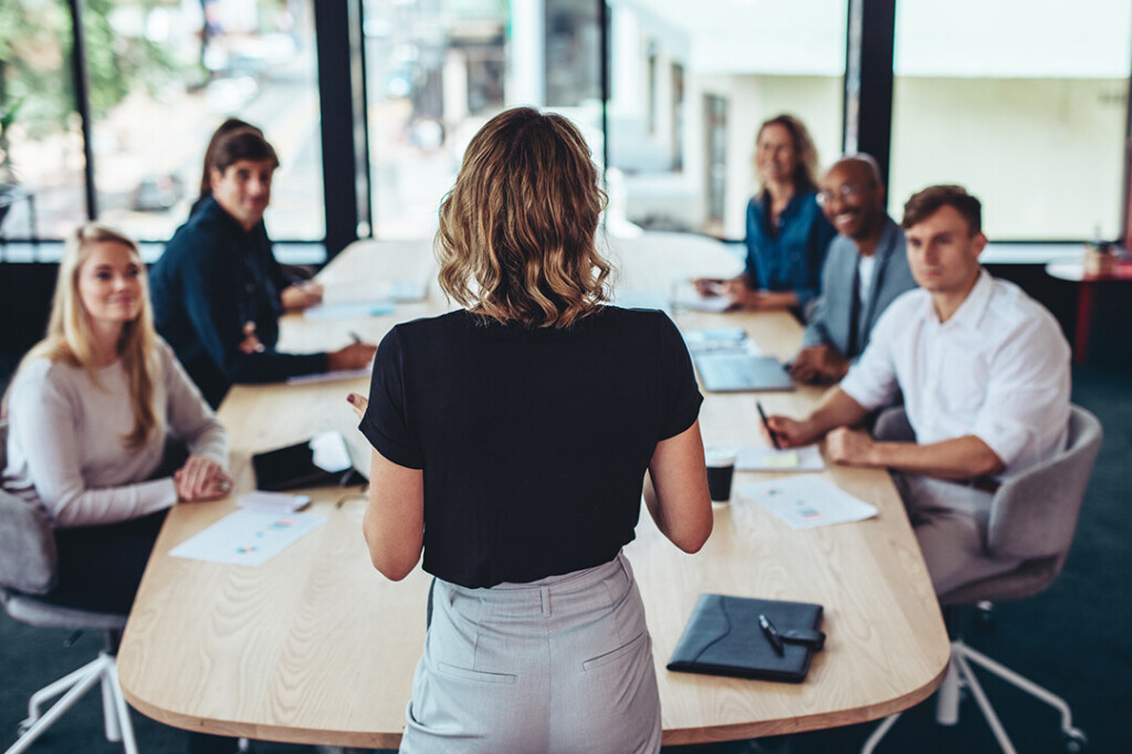 Frau steht vor einem Konferenztisch und spricht zu einer Gruppe von sechs Personen, die aufmerksam zuhören – modernes Business-Meeting in einem hellen Besprechungsraum.