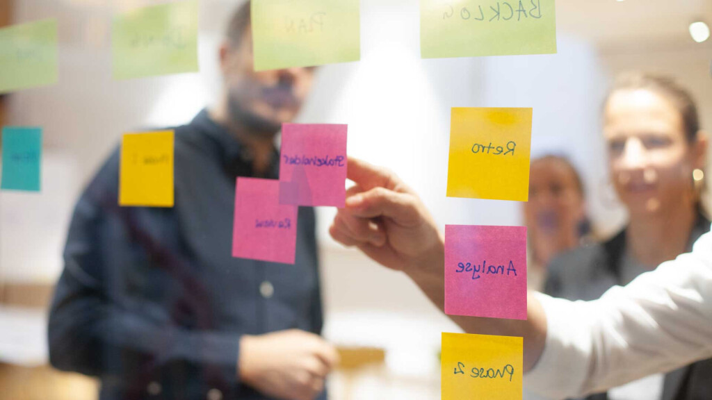 Teammitglieder betrachten bunte Haftnotizen an einer Glasscheibe während eines agilen Planungsmeetings oder Workshops.