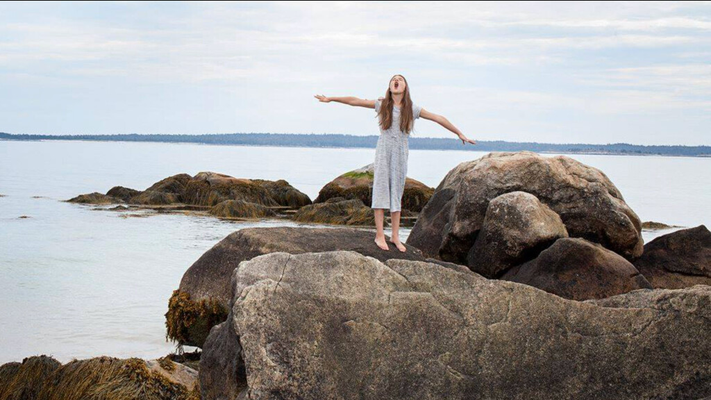 Mädchen steht barfuß auf Felsen am Meer, breitet die Arme weit aus und ruft in den Himmel; im Hintergrund ruhige See und ferne Küste.