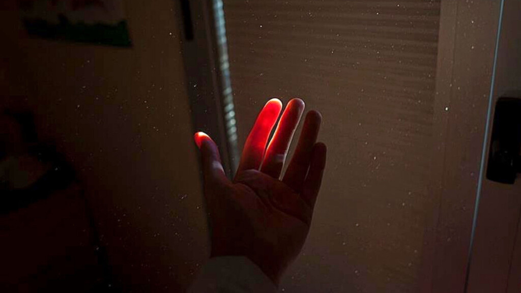 Hand in einem dunklen Raum, deren Finger von einem Lichtstrahl durchleuchtet werden, sodass sie rot leuchten.
